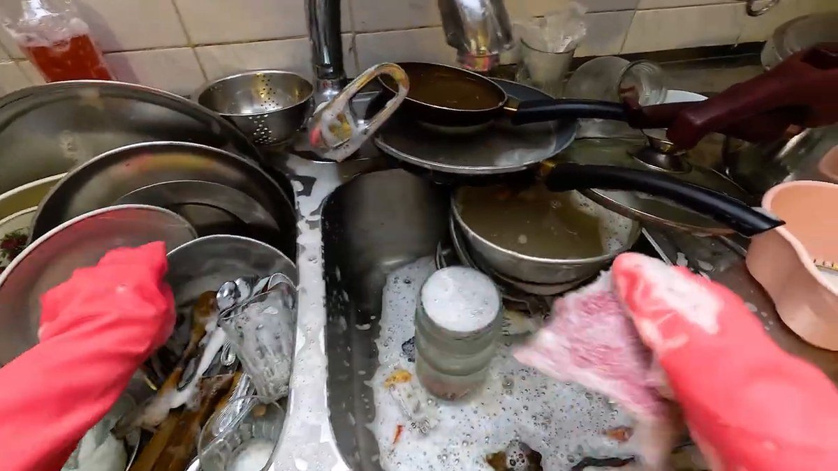 Mãos com luva rosa lavando louça numa pia cheia, gravadas em primeira pessoa pelo celular preso à cabeça