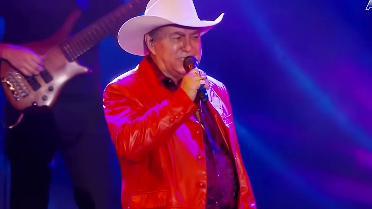 Milionário, de chapéu branco e jaqueta vermelha, cantando ao microfone num palco com luz azul