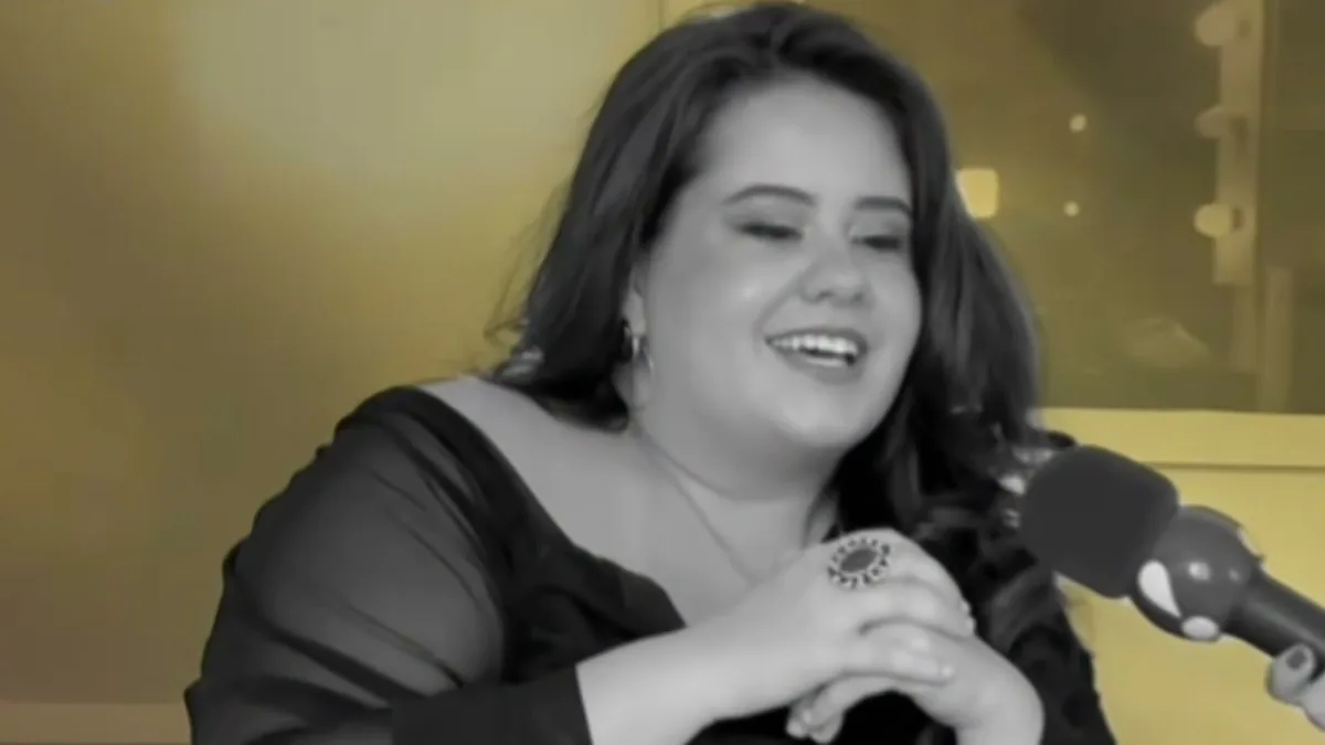 Karol Lannes, de blusa preta, sorri com as mãos juntas durante uma entrevista, em imagem preto e branco