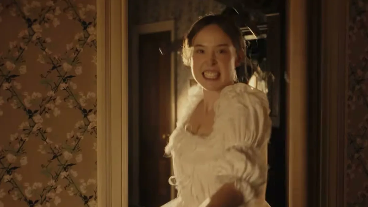 Ella Beatty como Lizzie Borden, de vestido branco, com expressão de fúria na porta de um quarto de papel de parede florido