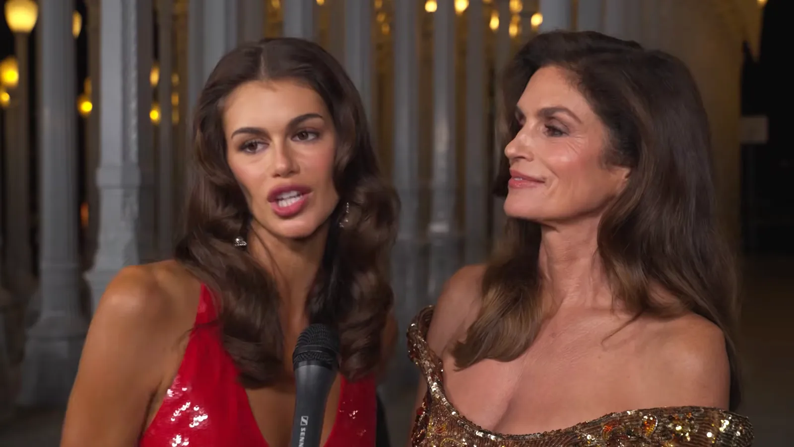 Kaia Gerber, de vestido vermelho, ao lado da mãe, Cindy Crawford, de vestido dourado, num evento à noite
