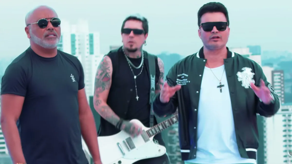Rick, de camiseta preta e óculos escuros, ao lado de um guitarrista e de Renner, de jaqueta verde, no alto de um prédio