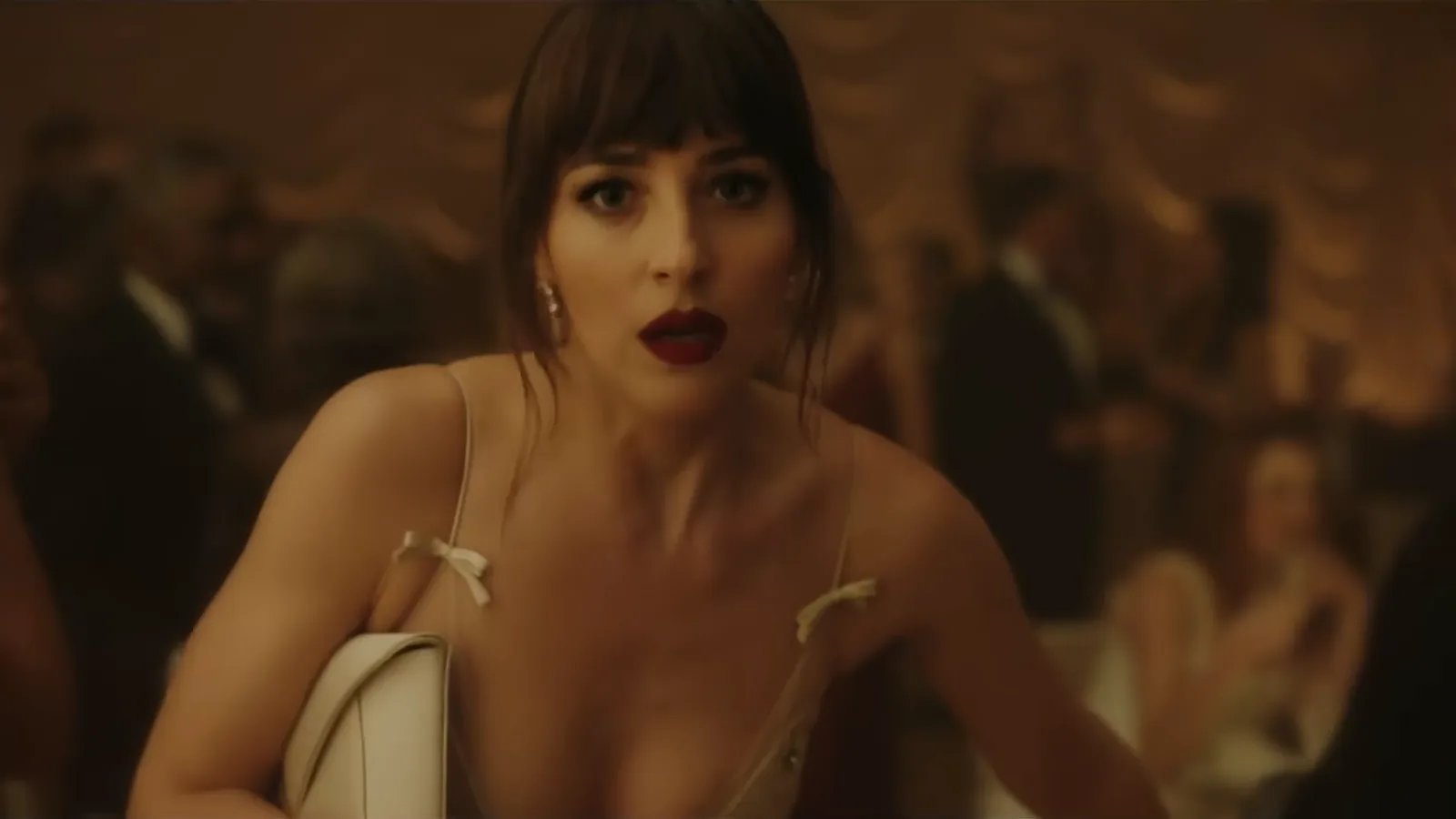 Dakota Johnson, de franja, batom vermelho e vestido claro, com expressão de espanto num salão de festa, em cena do clipe de Patient Zero