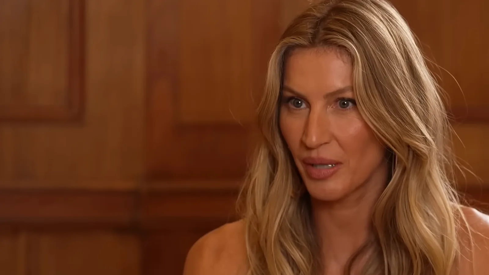 Gisele Bündchen, de cabelo loiro comprido, fala durante uma entrevista numa sala com paredes de madeira