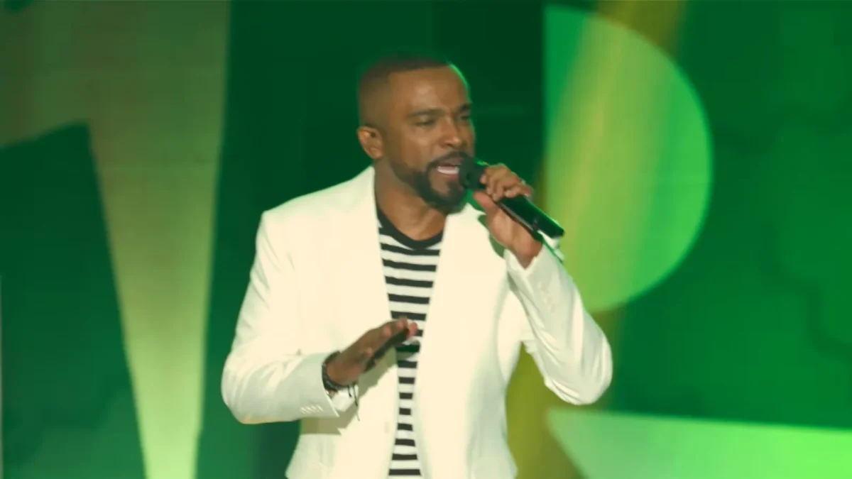 Alexandre Pires, de blazer branco e camisa listrada, canta ao microfone num palco com luz verde