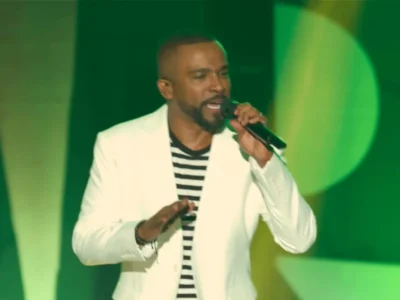 Alexandre Pires, de blazer branco e camisa listrada, canta ao microfone num palco com luz verde
