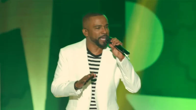 Alexandre Pires, de blazer branco e camisa listrada, canta ao microfone num palco com luz verde