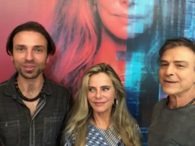 Kim Riccelli, Bruna Lombardi e Carlos Alberto Riccelli sorrindo lado a lado diante de um painel colorido