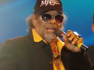 José Rico, de boné com o logo da dupla e óculos escuros, cantando ao microfone num palco com luz azul