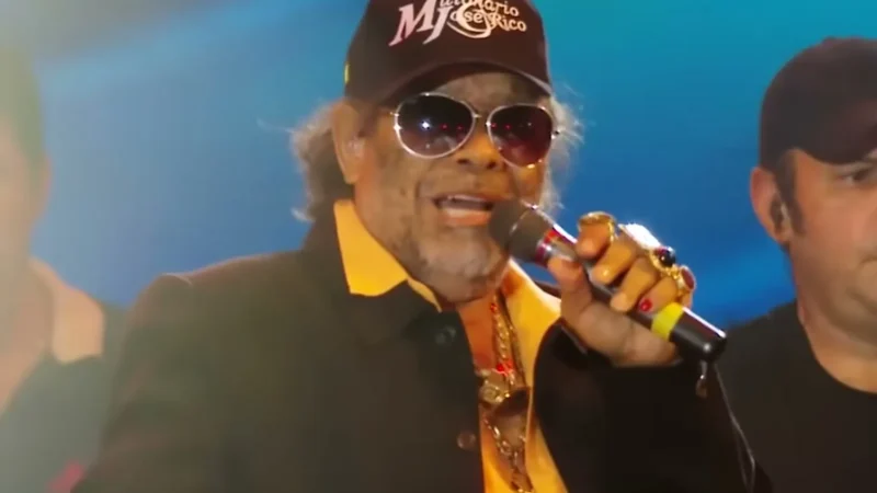 José Rico, de boné com o logo da dupla e óculos escuros, cantando ao microfone num palco com luz azul