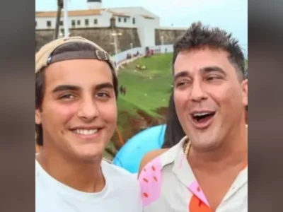 Marcelo Sangalo, de boné virado para trás e camiseta branca, sorri ao lado de André Marques, de camisa estampada, com o Farol da Barra ao fundo