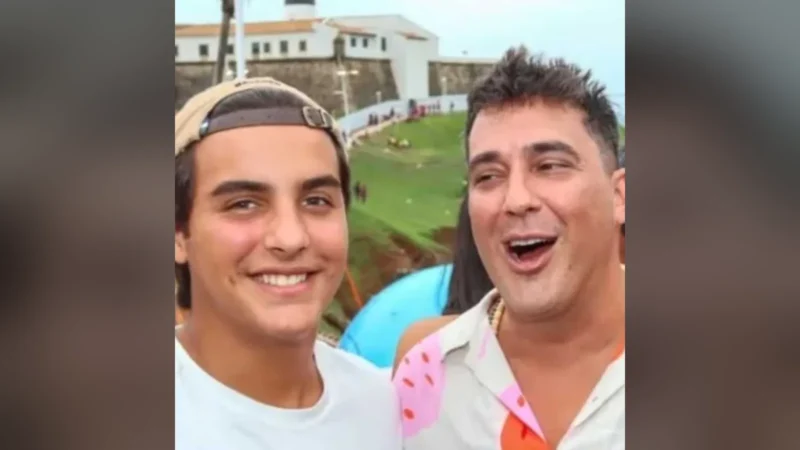 Marcelo Sangalo, de boné virado para trás e camiseta branca, sorri ao lado de André Marques, de camisa estampada, com o Farol da Barra ao fundo