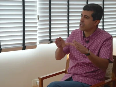 Marcius Melhem, de camisa estampada, sentado e gesticulando durante uma entrevista