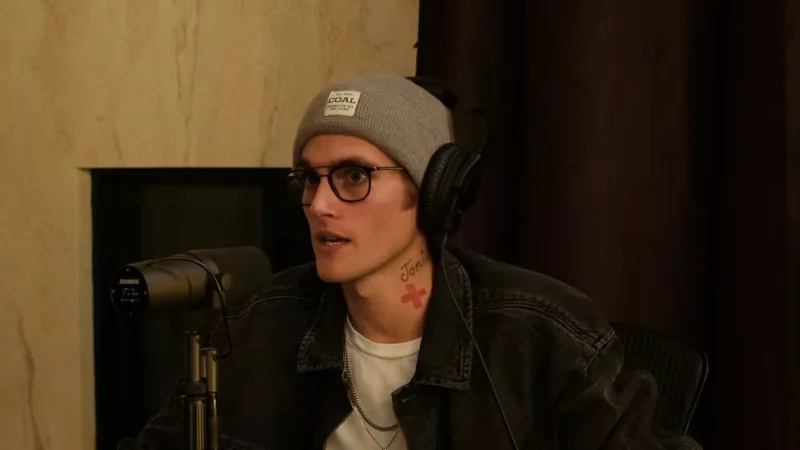 Presley Gerber, de gorro cinza, óculos de grau e fone de ouvido, fala num estúdio de podcast