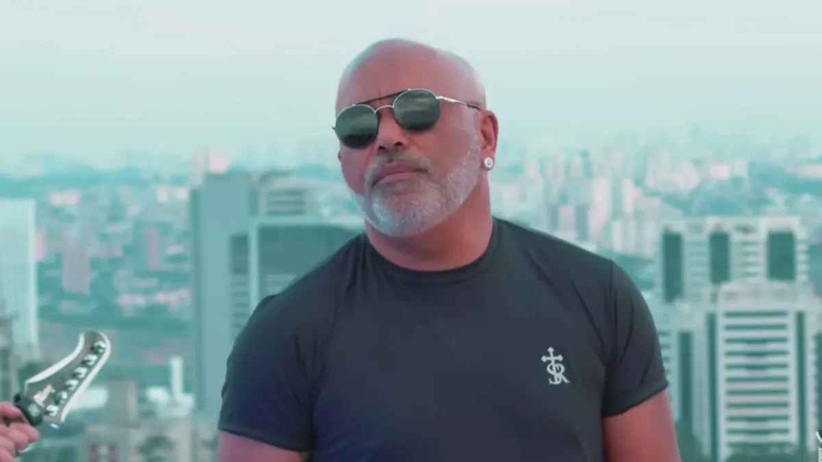 Rick, da dupla com Renner, de óculos escuros e barba branca, no alto de um prédio com a cidade ao fundo