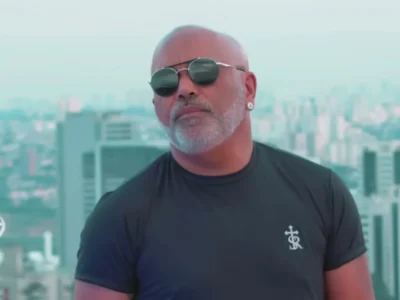 Rick, da dupla com Renner, de óculos escuros e barba branca, no alto de um prédio com a cidade ao fundo