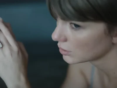 Taylor Swift, de franja e cabelo castanho, olha para baixo com a mão erguida e o anel de noivado à mostra, em cena do clipe de Patient Zero