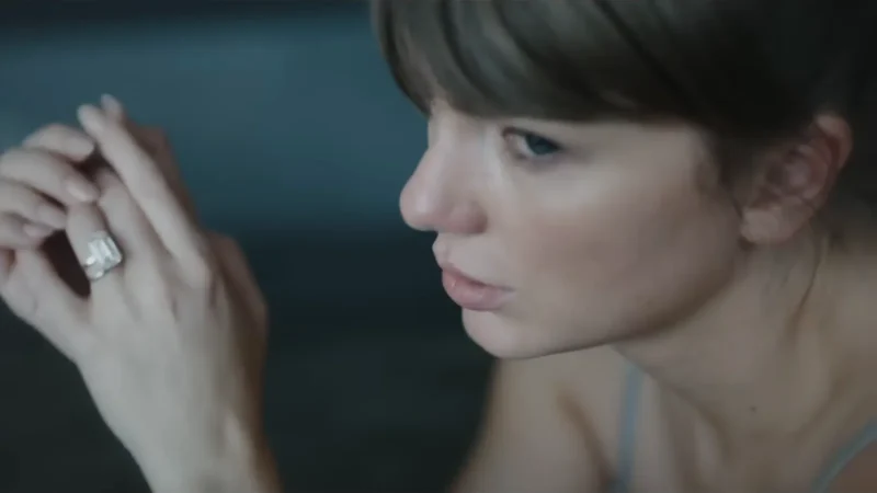 Taylor Swift, de franja e cabelo castanho, olha para baixo com a mão erguida e o anel de noivado à mostra, em cena do clipe de Patient Zero